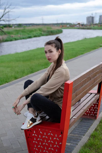 Ελκυστική μελαχρινή γυναίκα κάθεται σε ένα παγκάκι στο πάρκο. ρούχα μόδας, κομψή εμφάνιση — Φωτογραφία Αρχείου