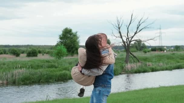 Een gelukkige blanke man die een brunette in zijn armen houdt en haar ronddraait op een zomerdag. Paar, date, liefde en lifestyle concept. Een jongeman omcirkelt zijn vriendin. slow motion — Stockvideo