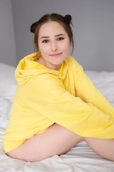 快乐的 积极的黑发女人 穿着黄色的连帽衫 躺在白色的卧室里 床上躺着白色的亚麻布 快乐的人千年一代 时髦的青少年 — 图库照片