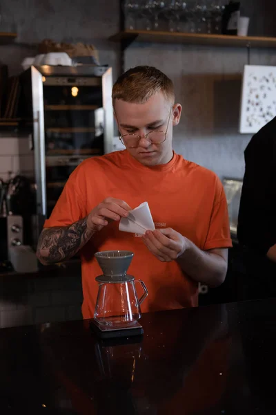 blonde guy in orange t-shirt preparing Alternative coffee brewing: chemex in modern loft coffee shop