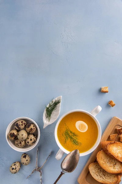 Bouillon, quail egg and fennel leaf in white soup bowl on blue background. Surrounded by toasted bread, croutons, tongs, spoon and eggs. Vertical orientation. Healthy eating and dieting. Copy space. — Stock Image Bouillon Quail Egg Fennel Leaf White Soup Bowl Blue Background Royalty Free Stock Images