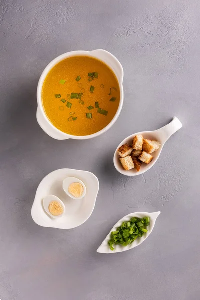 Bouillon in white soup bowl, chopped spring onions or scallions, quail eggs and croutons on gray stone background. Table top view. Vertical orientation. Homemade healthy eating and dieting concept. — Stock Image Bouillon White Soup Bowl Chopped Spring Onions Scallions Quail Eggs Stock Photo