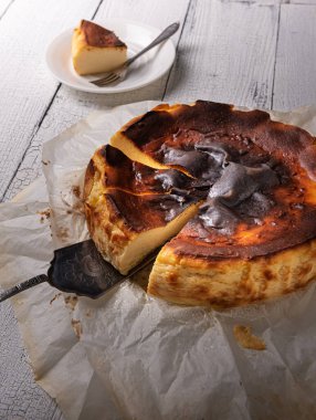 Beyaz ahşap masa üzerinde hamur işi kağıt içinde gümüş servis spatula üzerinde kesilmiş basklı yakılmış cheesecake. Arka planda plaka üzerine odaklanmamış kısım. Ev yapımı İspanyol tatlısı, köy usulü..