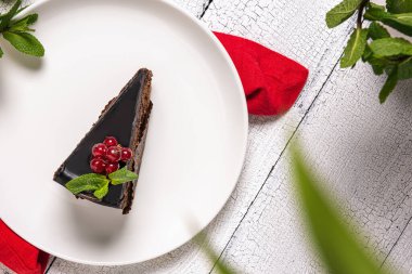 Mozart adında bir dilim çikolatalı kek üzerine kırmızı üzümlü üzümler ve beyaz tabakta nane yaprağı, kırmızı keten masa örtüsü ve beyaz ahşap masa örtüsü ile süslenmiş. Odaklanmamış bitki yaprakları ön planda.