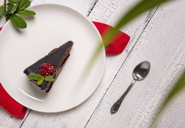 Mozart adında bir parça çikolatalı kek üzerine kırmızı frenk üzümü ve nane yaprağı ile süslenmiş beyaz tabak, kırmızı masa örtüsü ve beyaz ahşap masa örtüsü. Odaklanmamış bitki yaprakları ön planda.