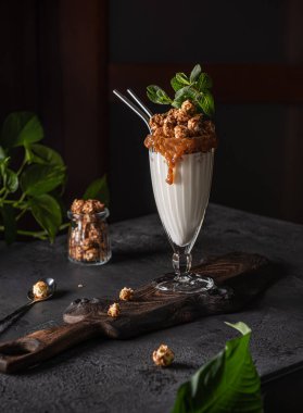 Büyük bardakta patlamış mısır, karamel, nane ve tahta tahtada duran iki metal pipetle ve doğal bitkilerle süslenmiş siyah masanın üzerinde duran lezzetli büyük boy milkshake ile hayat. Seçici odak.