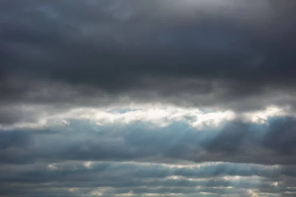 Beautiful sky landscape background with stripes of heavy dark gray, almost black stormy clouds. Textured clearly visible sun rays through gaps. Some clouds are litted with sun behind them.