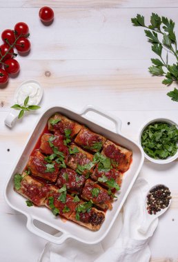 Domates soslu lahana ruloları, kiraz domatesleri, maydanoz, ekşi krema ve keten peçeteyle bembeyaz bir tavayla dümdüz yatıyordu. Dikey beyaz ve bej doğal ahşap arka plan. Boşluğu kopyala.