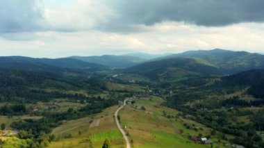 Havadan panoramik manzara. Dağların eteklerinde toprak yol uzanıyor. Yeşillikler arasında tepelerin yamaçlarındaki evler. Dağlar kozalaklı ormanlarla kaplı, dağların yamaçlarında yeşil çimenler, temiz oksijen zengini hava. Ukrayna Karpatları