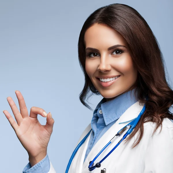 Mujer joven médico mostrando signo de mano bien, en gris —  Fotos de Stock