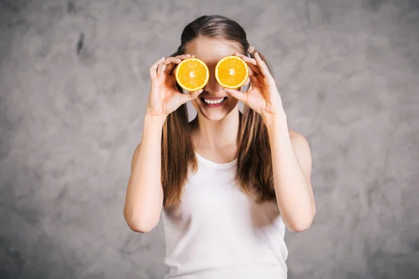 Smiling young woman covering eyes with orange halves on concrete background — Stock Photo, Image