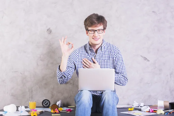 Lächelnder kaukasischer Mann mit Laptop sitzt auf unordentlichem Desktop und zeigt ok Zeichen — Stockfoto