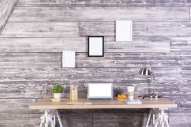 Modern tasarımcı çalışma alanı boş beyaz dizüstü, kahve, lamba, çeşitli ileti örneği öğeleri ve yukarıda ahşap duvarda asılı üç boş resim çerçeveleri ile önden görünümü.