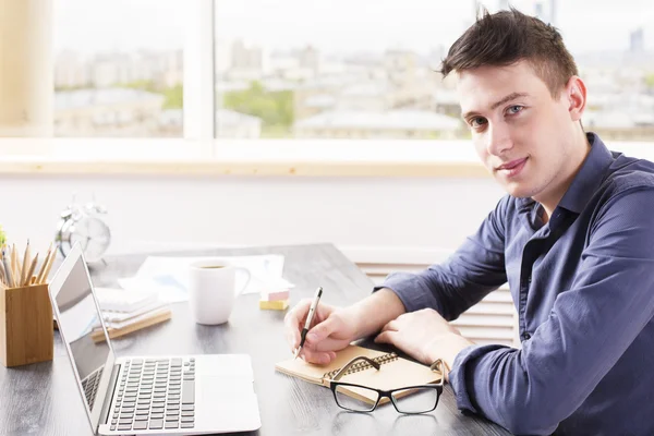 Концепция образования: молодой человек пишет в блокноте на рабочем столе с ноутбуком, кофейной чашкой, очками и другими предметами — стоковое фото