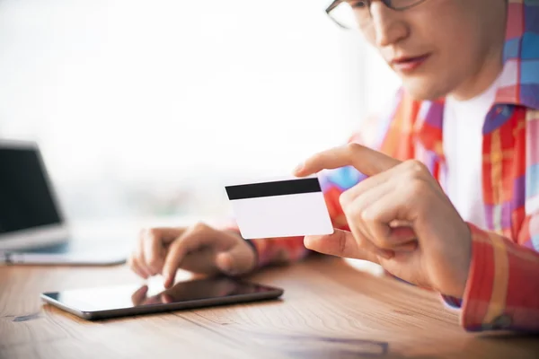 Concepto de compras en línea con el hombre joven casual que sostiene la tarjeta de crédito y el uso de la tableta en escritorio de madera —  Fotos de Stock