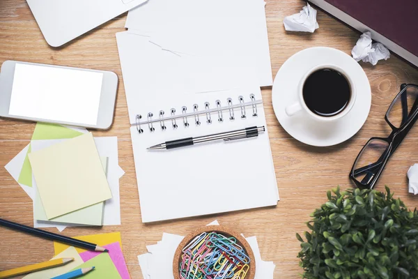 Lugar de trabajo desordenado con teléfono inteligente blanco en blanco, bloc de notas en espiral abierto, taza de café, vasos, planta y varios artículos de papelería. Prepárate. —  Fotos de Stock