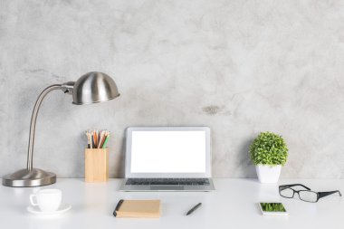 Boş beyaz dizüstü bilgisayar, akıllı telefon, gözlük, not defteri, kahve fincanı, renkli kalemler, bitki ve beton duvar arka plan üzerinde masa lambası ile Yaratıcı tasarımcı masaüstü. Alay etmek