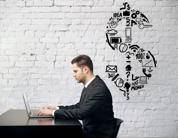 Side view of businessman using laptop at desk placed against white brick wall with dollar sign sketch. Financial growth concept — Stock Photo, Image