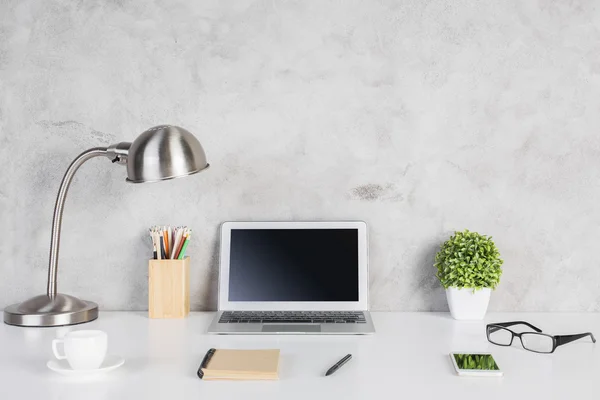 Creative designer desktop with blank laptop computer, smartphone, glasses, notepad, coffee cup, colorful pencils, plant and table lamp on concrete wall background. Mock up