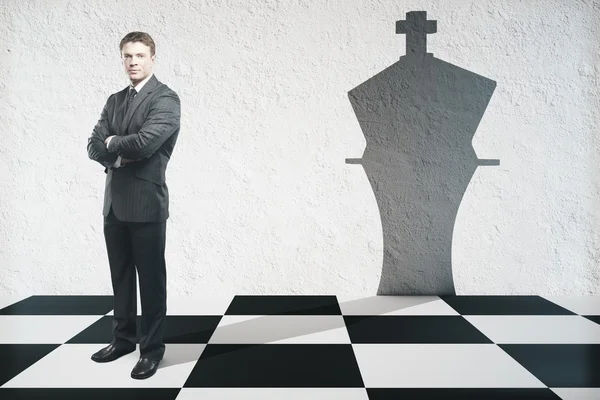Empresário com braços dobrados em pé no tabuleiro de xadrez com sombra rei peça na parede de concreto texturizado. Conceito de liderança — Fotografia de Stock
