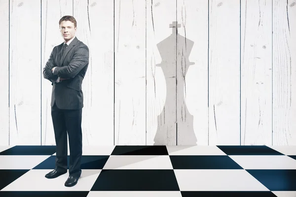 Businessman with folded arms standing on chessboard with king piece shadow on textured wooden wall. Leadership concept — Stock Photo, Image