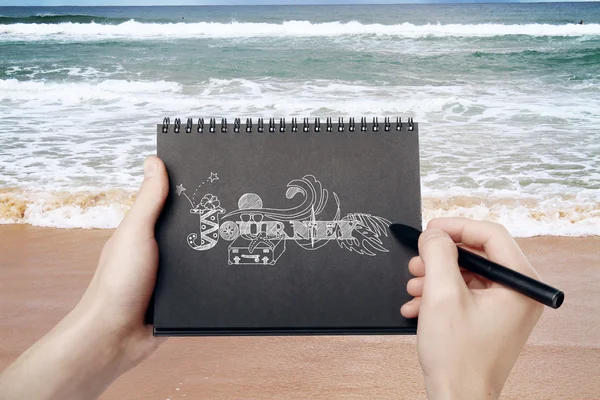 Tangan laki-laki memegang notepad spiral hitam dan menggambar sketsa perjalanan di latar pantai. Konsep perjalanan — Stok Foto
