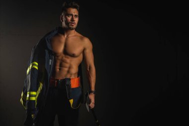 young handsome adult, muscular firefighter in uniform holding ax of fire equipment in his hands, pensive, isolated on dark background. Low key. Protection concept. there is a place for an inscription, the text clipart