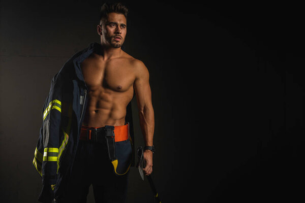 young handsome adult, muscular firefighter in uniform holding ax of fire equipment in his hands, pensive, isolated on dark background. Low key. Protection concept. there is a place for an inscription, the text