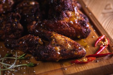 İştah açıcı, ızgarada pişirilmiş altın tavuk kanatları ahşap bir tahtada servis ediliyor, arka planda taze yeşillikler, üst manzara. Koyu arkaplan, düşük tuş