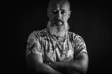 Ciddi, kel, sakallı, tişörtlü bir adamın portresi. Kolları önünde çaprazlanmış. Koyu bir arka planda poz veren gür sakallı. Moda ve tarz. Kapatın, yazı için boşluk