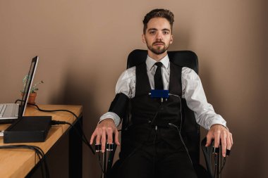 Korkmuş, endişeli, dalgın, sandalyede oturan, yalan makinesinde test yapan, yalan makinesi sensörleri takan, gri tuğlalardan oluşan bir arka planda oturan, düz duran, tepeden bakan bir adam.