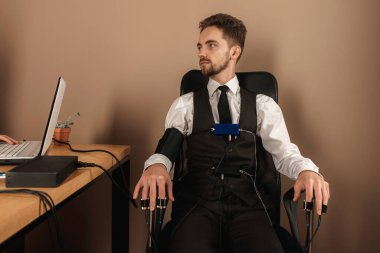 Odaklanmış, sakallı düşünceli bir adam parlak bir odada yalan makinesini test ediyor. Sandalyede otururken parmaklarına yalan makinesi yerleştirmek için sensör takıyor. Doğruluk kavramı, yalan makinesi.