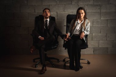 Koyu renk takım elbiseli, beyaz gömlekli genç bir adam ve koyu deri bir ofis sandalyesinde yan yana oturan güzel bir kız. Kız adamın elini tutuyor ve gri bir arka planda onu incitiyor. Alçak tuş