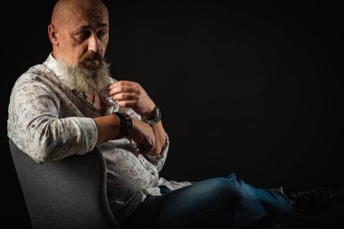 Yakın plan, duygusal, düşünceli, yakışıklı, sakallı genç bir iş adamının portresi. Sandalyeye oturmuş, elleriyle sakalını okşuyor. Alçak tuş koyu arkaplanda izole edildi. Vücut dili