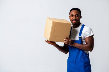 Karton kutulu bir kurye, mavi tulumlu genç bir Afro-Amerikan adam ve elinde kağıt kutu tutan beyaz bir tişört. Beyaz bir arka planda izole edilmiş. Teslimat, posta, sevkiyat
