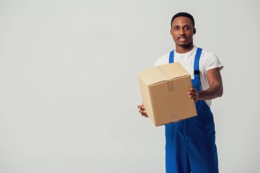 Karton kutulu bir kurye, mavi tulumlu genç bir Afro-Amerikan adam ve elinde kağıt kutu tutan beyaz bir tişört. Beyaz bir arka planda izole edilmiş. Teslimat, posta, sevkiyat