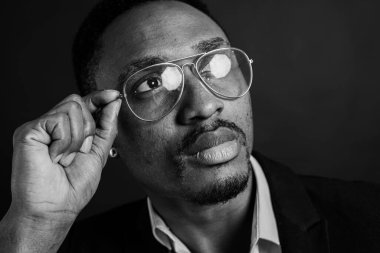 Çekici, yakışıklı, şık Afro-Amerikan iş adamının şık takım elbiseli ve beyaz gömlekli portresi gözlükleri elle ayarlar, koyu arka planda izole eder. Alçakgönüllülük. Seçici odak