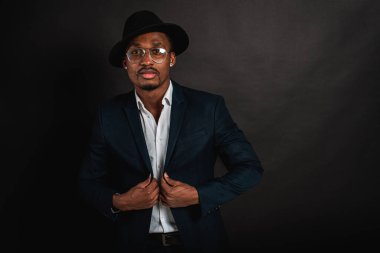 Çekici, yakışıklı, güzel, ciddi ve stil sahibi Afro-Amerikan iş adamının portresi şık takım elbiseli, beyaz gömlekli ve siyah şapkalı koyu arka planda. Alçakgönüllülük. Seçici odak