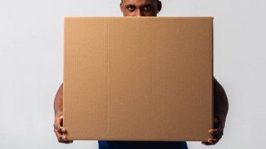 Teslimatçı elinde beyaz tişört ve elinde kutu olan mavi pantolon tutuyor. Beyaz bir arka planda izole edilmiş. Teslimat, posta, sevkiyat, yükleyici, kurye. Kutuyu kapat