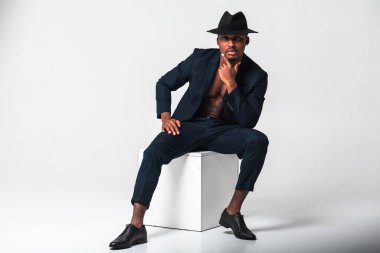 Çekici, yakışıklı, güzel ve şık bir iş adamının portresi şık bir takım elbise ve siyah şapkalı Afro-Amerikan modeli beyaz bir arka planda izole bir şekilde beyaz bir meydanda oturuyor. Yüksek tuş.