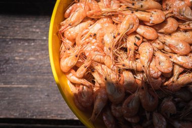 Haşlanmış karides, gri-kahverengi ahşap bir yüzey üzerinde sarı bir kâse içinde soyulmamış, açık renk gölge, güneş ışığı. Kasede bir sürü karides var. Karidese yakın çekim. Metin için boşluk olan üst görünüm. Pankart