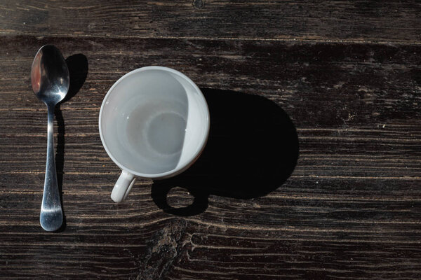 Cup of coffee in a white cup and a metal teaspoon on a gray-cinnamon wooden surface, light shadow, sunlight of morning coffee, top view