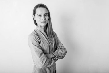 Açık gri elbiseli, mavi gözlü, sarı saçlı ve yüzünde bir gülümseme olan kendinden emin bir kadın kollarını kavuşturmuş beyaz duvarın yanında duran kameraya bakıyor. İnsanlar, gençlik, güzellik, yaşam tarzı konsepti