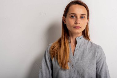 Açık gri elbiseli, mavi gözlü, sarı saçlı ve yüzünde bir gülümseme olan kendinden emin bir kadın kollarını kavuşturmuş beyaz duvarın yanında duran kameraya bakıyor. İnsanlar, gençlik, güzellik, yaşam tarzı konsepti 