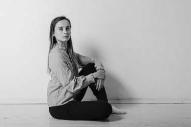 Siyah ve beyaz bir fotoğraf, yerde bacakları çapraz oturan, gri gömlekli ve siyah pantolonlu, düşünceli, üzgün, üzgün parlak bir odada duran genç bir kız. Depresyon ve yalnızlık
