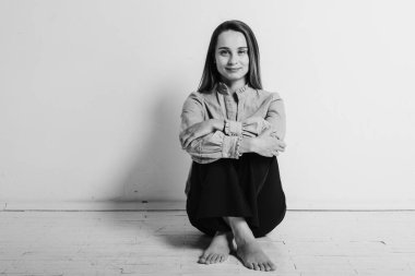 Siyah ve beyaz bir fotoğraf, yerde bacakları çapraz oturan, gri gömlekli ve siyah pantolonlu, düşünceli, üzgün, üzgün parlak bir odada duran genç bir kız. Depresyon ve yalnızlık