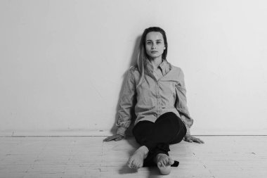 Yerde oturan genç ve güzel bir kız gri gömlekli ve siyah pantolonlu kameraya bakarak bacaklarını çaprazladı. Düşünceli, üzgün, parlak bir odada üzgün. Depresyon ve yalnızlık