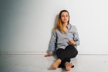 Yerde oturan genç ve güzel bir kız gri gömlekli ve siyah pantolonlu kameraya bakarak bacaklarını çaprazladı. Düşünceli, üzgün, parlak bir odada üzgün. Depresyon ve yalnızlık