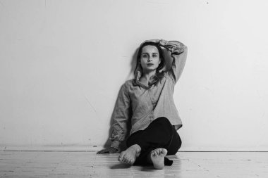 Siyah ve beyaz bir fotoğraf, yerde bacakları çapraz oturan, gri gömlekli ve siyah pantolonlu, düşünceli, üzgün, üzgün parlak bir odada duran genç bir kız. Depresyon ve yalnızlık