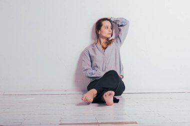 Yerde oturan genç ve güzel bir kız gri gömlekli ve siyah pantolonlu kameraya bakarak bacaklarını çaprazladı. Düşünceli, üzgün, parlak bir odada üzgün. Depresyon ve yalnızlık